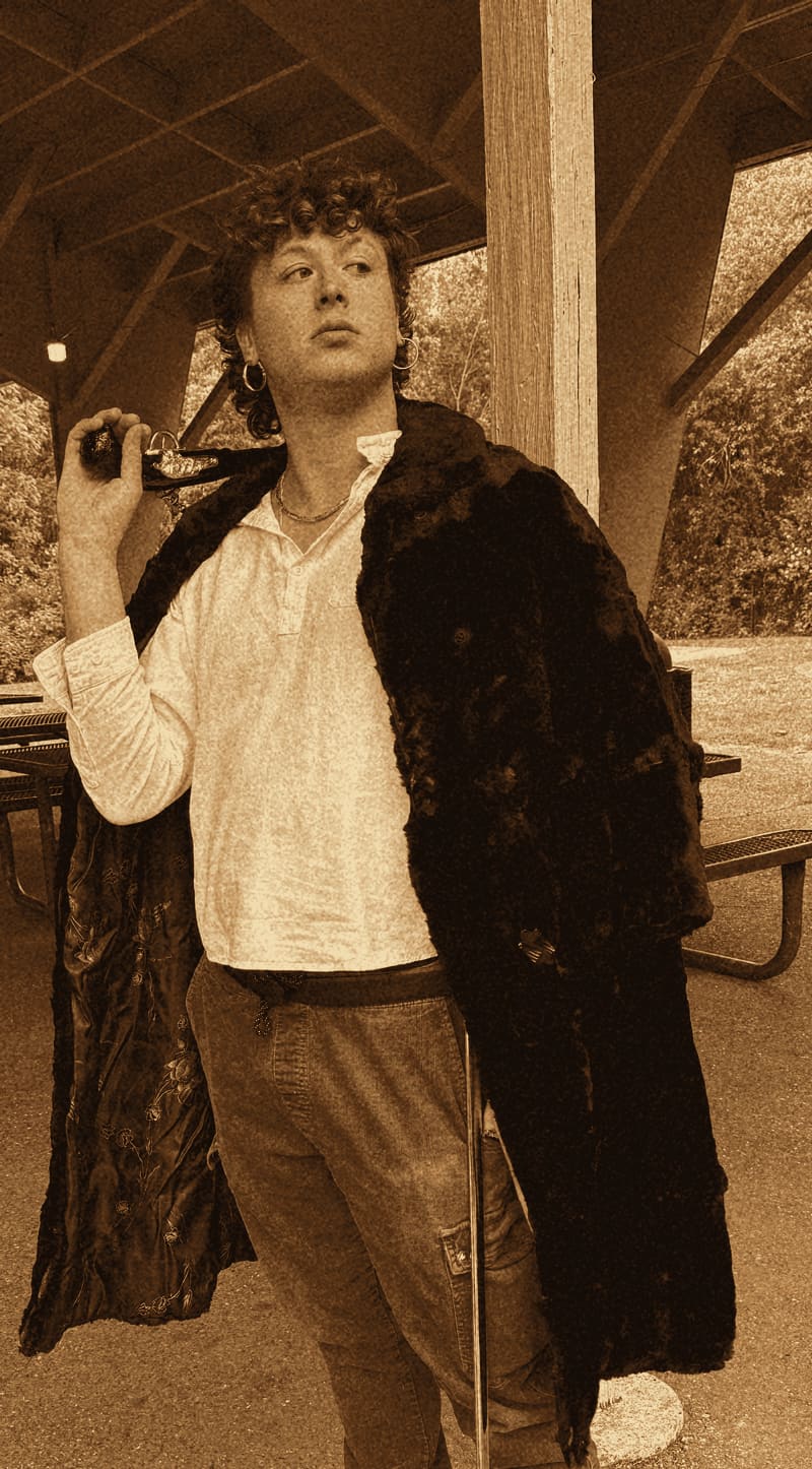 Exhibit T: the suspect posing heroically under a park pavilion in a full-length black fur coat over a white peasant shirt, rust corduroy cargo pants and black Doc Martens, a silver-handled cane over his shoulder and a hoop earring, gazing into the middle distance.