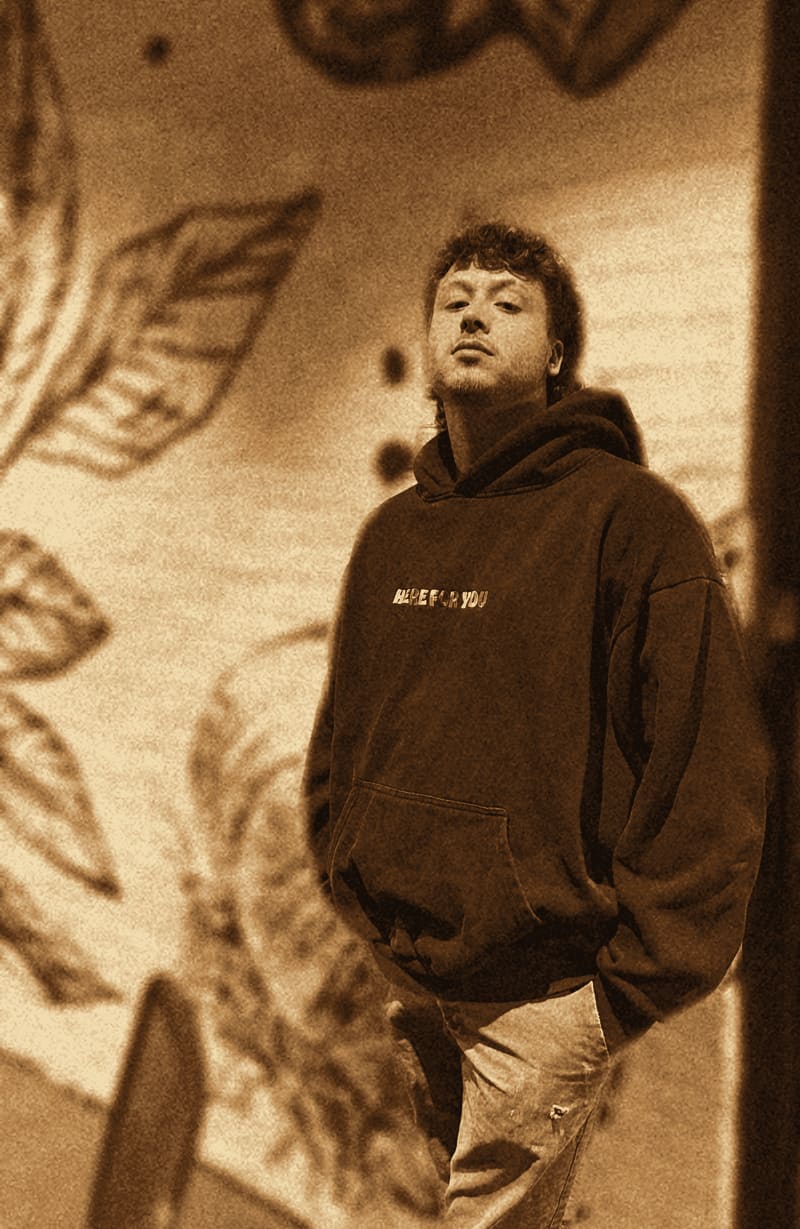 Exhibit G: the suspect leaning against a white brick wall painted with black ink leaves, wearing a navy hoodie that says HERE FOR YOU in rainbow letters, hands in his pockets, giving the camera a look he has clearly practiced.