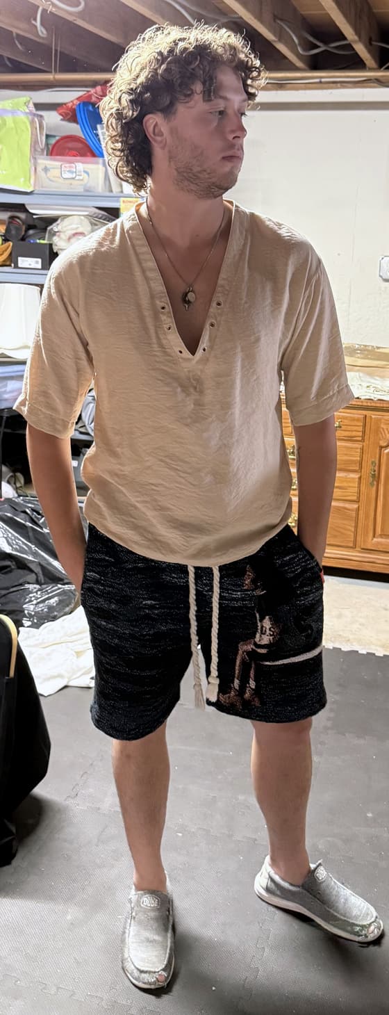 The suspect, photographed head to toe in a basement: curly hair, a deep-V linen shirt, a shark-tooth necklace, drawstring shorts and slip-on shoes. Staring off into the distance like he's somewhere better.