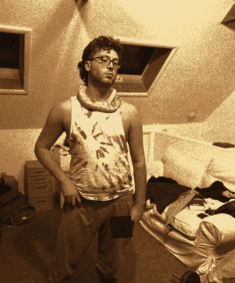 Exhibit B: the suspect in an attic bedroom wearing glasses, a tie-dye muscle tank and grey sweatpants, one hand on his hip, with a live yellow python draped around his neck like a scarf. A black bar covers the writing on his sweatpants.